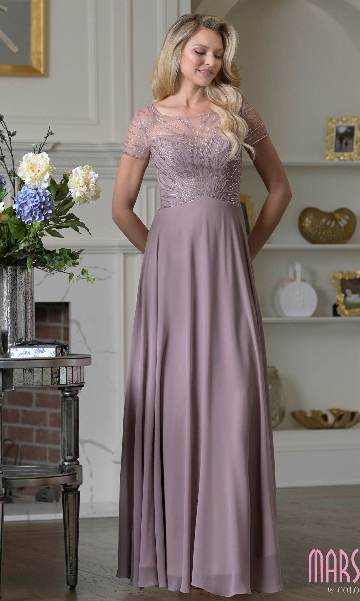 A woman in a Marsoni M351 Long Formal Dress, dusty rose chiffon with sheer short sleeves and an A-line, floor-length silhouette, stands indoors beside a table topped with flowers.